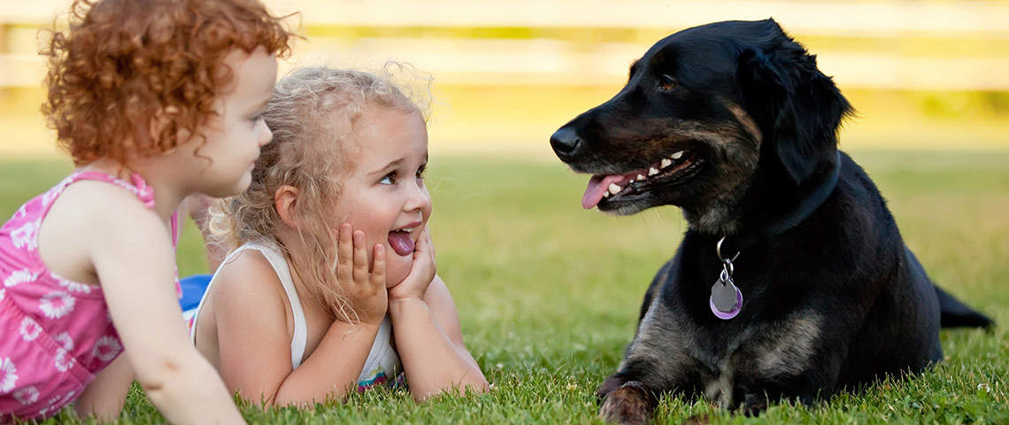 Two girls and a black dog laying on grass.
